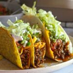 Two crunchy beef tacos filled with seasoned ground meat, shredded lettuce, cheese, guacamole, and sour cream on a plate.