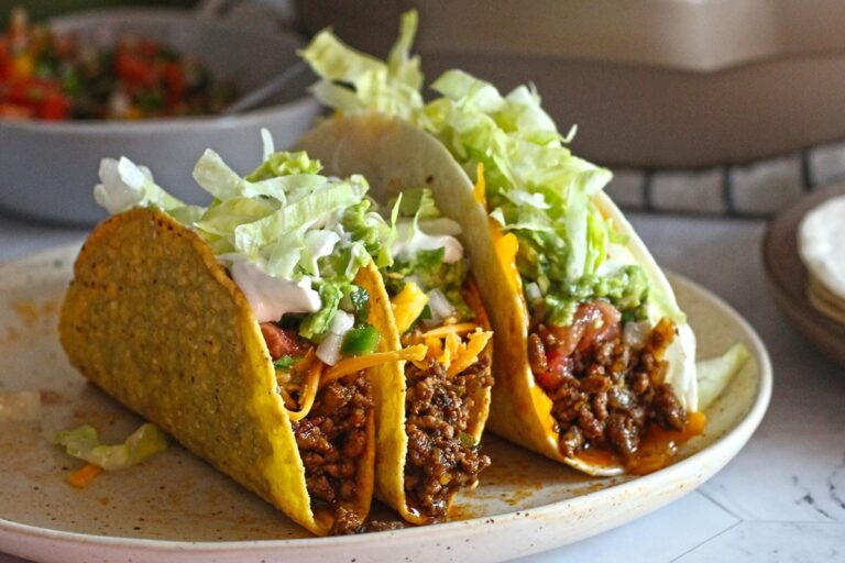Two crunchy beef tacos filled with seasoned ground meat, shredded lettuce, cheese, guacamole, and sour cream on a plate.