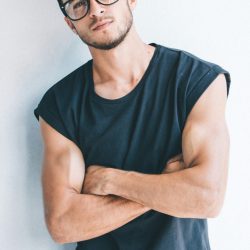 mubariz-mehdizadeh-Py8F6-hRn5o-unsplash A man with glasses crossing his arms, wearing a black sleeveless shirt, and giving a serious expression against a plain white wall.